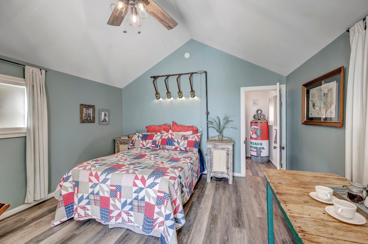 A cozy bedroom features a queen-size bed with a decorative quilt in red, white, and blue patterns. The room is illuminated by a ceiling fan with light fixtures, while a nightstand and a table with cups are positioned nearby. Light green walls and wood flooring enhance the inviting atmosphere.