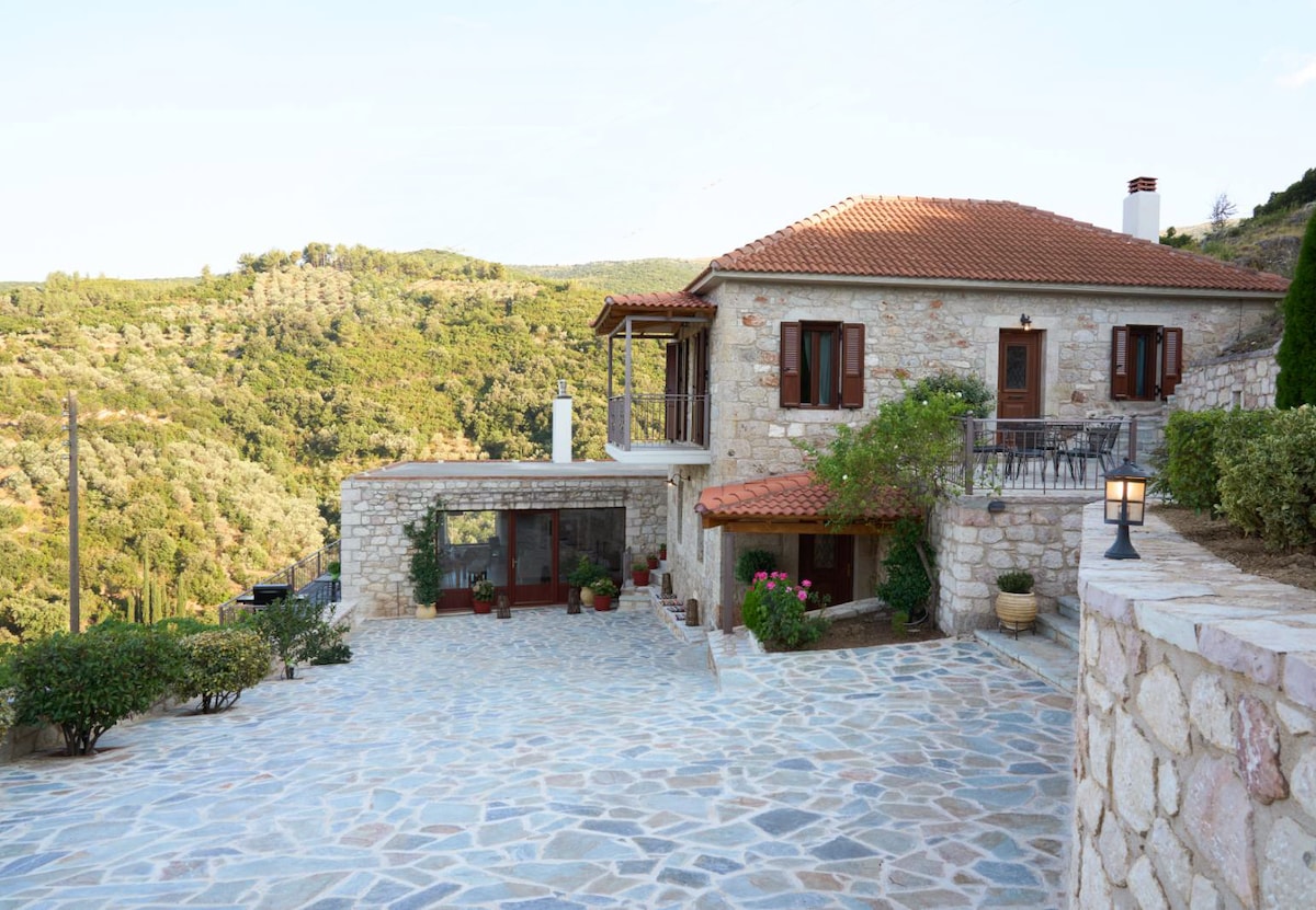 A traditional stone villa is positioned amidst lush green hills, showcasing a spacious stone-paved courtyard. The building features a red-tiled roof, wooden shutters, and an inviting balcony area. Surrounding gardens enhance the property's natural charm, creating a serene outdoor atmosphere.