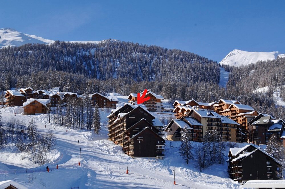 A scenic view of a ski resort nestled among snow-covered mountains. The image showcases several chalet-style buildings, blending harmoniously with the winter landscape, while ski slopes are visible in the foreground, indicating direct access to skiing activities.