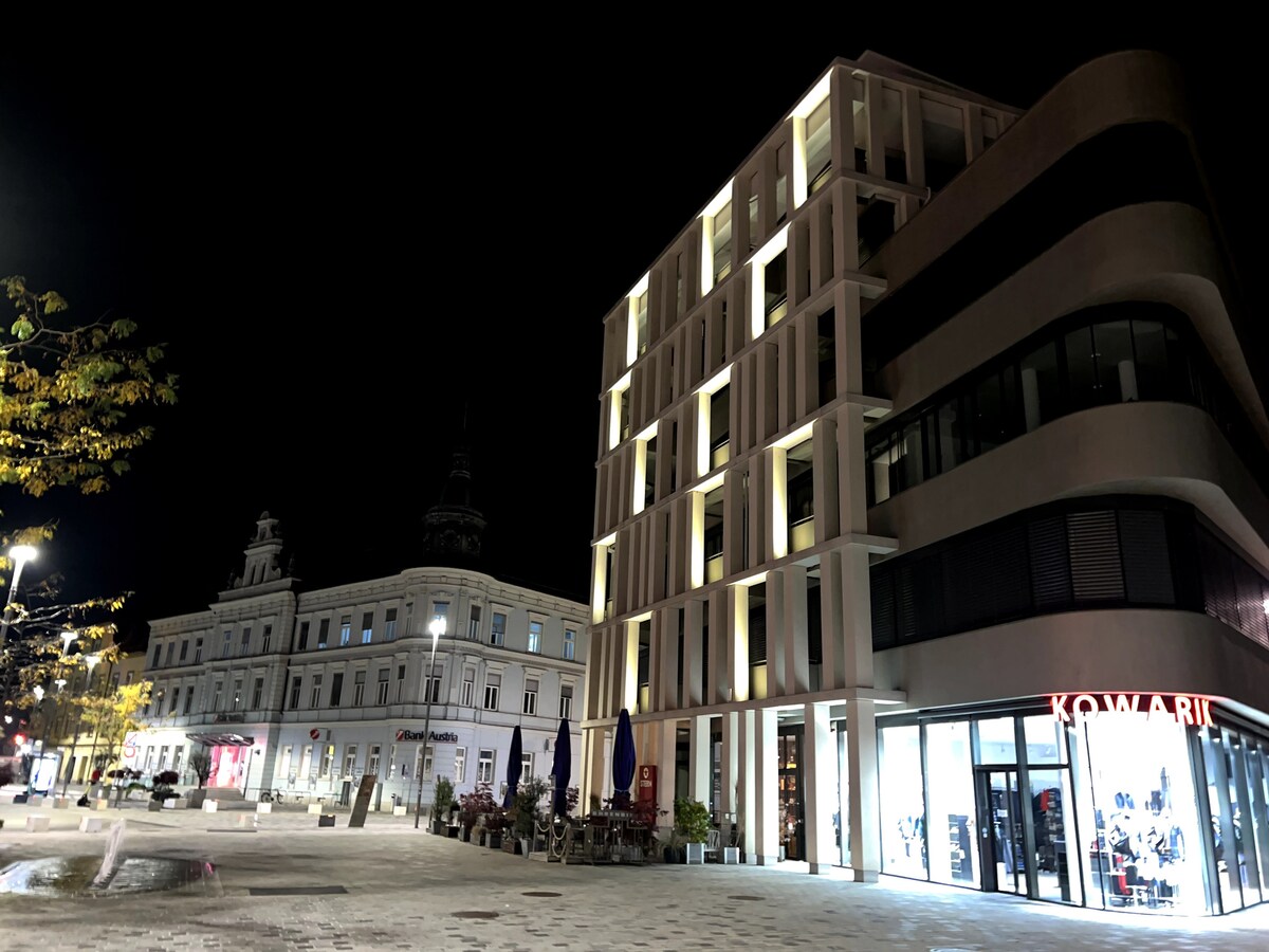 A modern building is illuminated at night, showcasing sleek architectural lines. Nearby, historic structures can be seen, featuring decorative facades. The square is designed with paved walkways and scattered seating areas, surrounded by trees and lighting that enhances the urban environment.