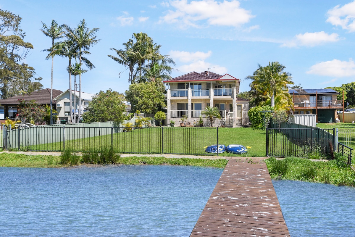 A two-story waterfront home is visible, framed by a neatly manicured lawn and tropical palm trees. A wooden jetty extends into the water, with moored water sports equipment nearby. The exterior features a mix of light-colored walls and a red-tiled roof.