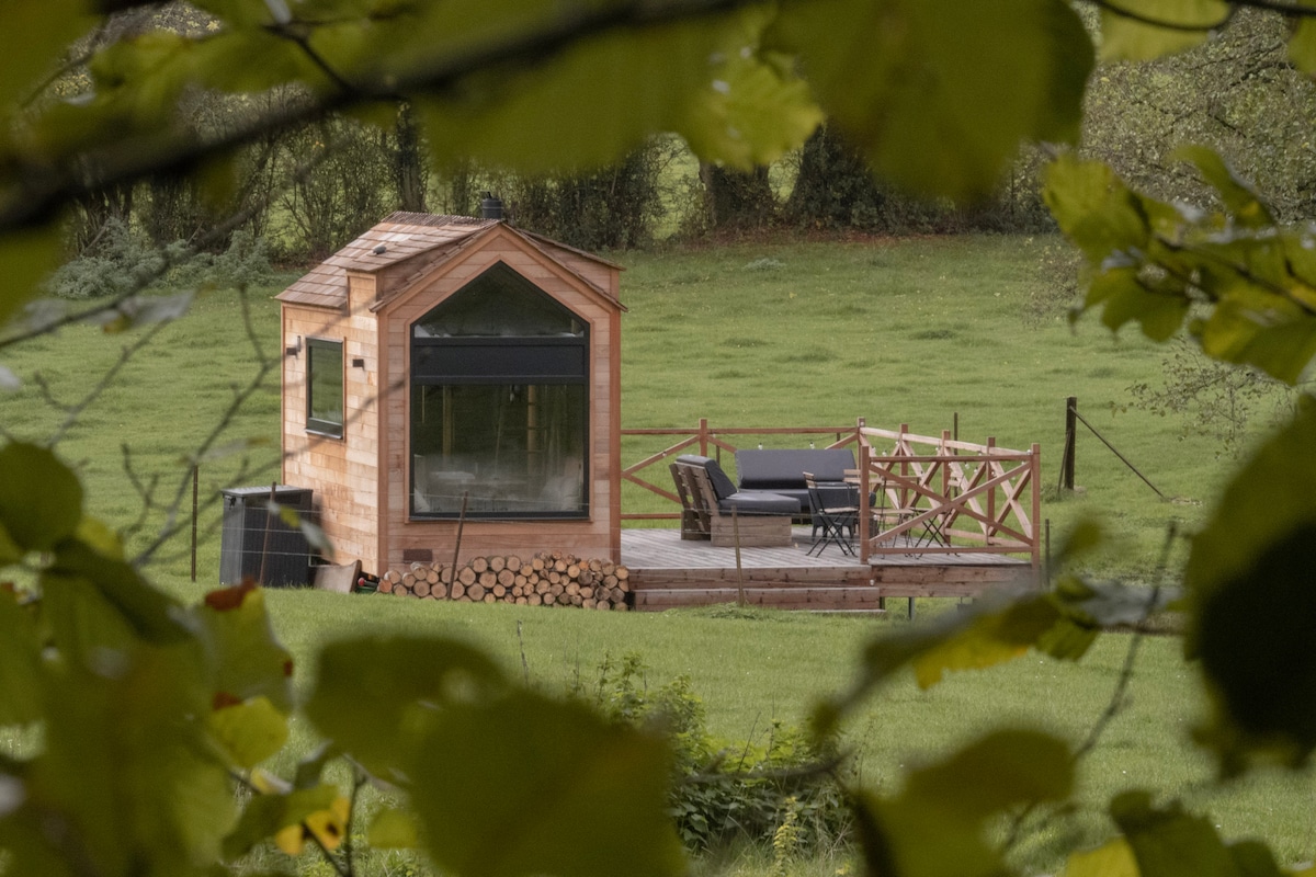 The Tiny Michou is framed by lush greenery, showcasing its wooden exterior and large window. A spacious deck features a seating area, enhancing the serene outdoor setting. Firewood is neatly stacked nearby, surrounded by a peaceful landscape.