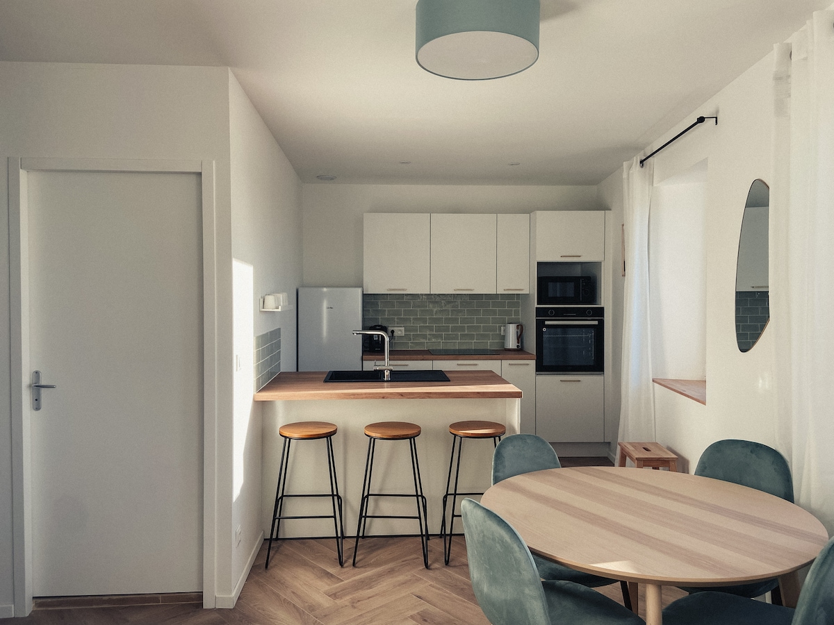 A modern kitchen space features a central island with three wooden bar stools. Sleek cabinetry and stainless steel appliances are visible. A round dining table with light green chairs is positioned nearby, complemented by natural light from a window.