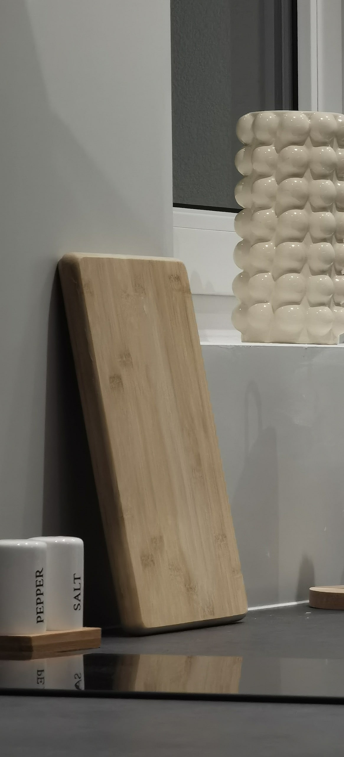 A wooden cutting board leans against the wall, showcasing a natural finish. To its right, a sculptural white vase with a textured surface adds a touch of contemporary design. Two canisters labeled 'SALT' and 'PEPPER' sit on a wooden tray, contributing to a functional kitchen space.