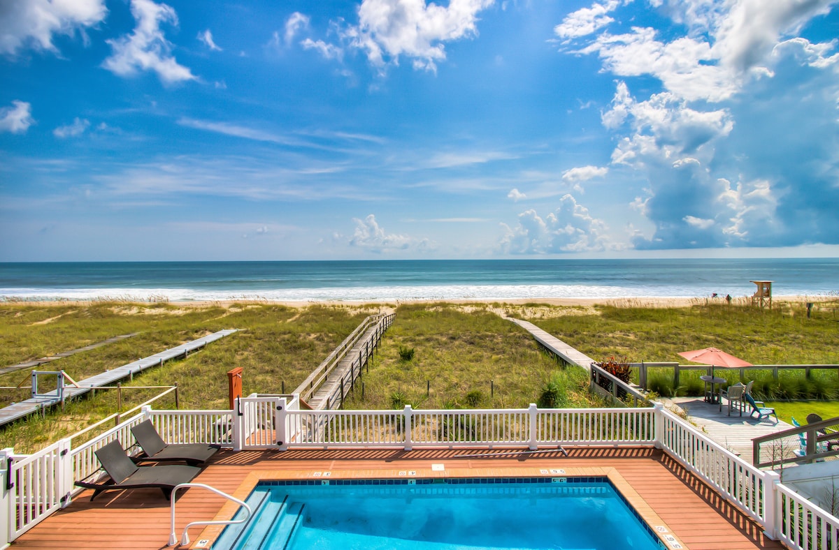 A private pool is highlighted in the foreground, bordered by a spacious deck with lounge chairs. Beyond, sandy paths lead to the beach, where gentle waves meet the shore under a bright blue sky dotted with soft clouds.