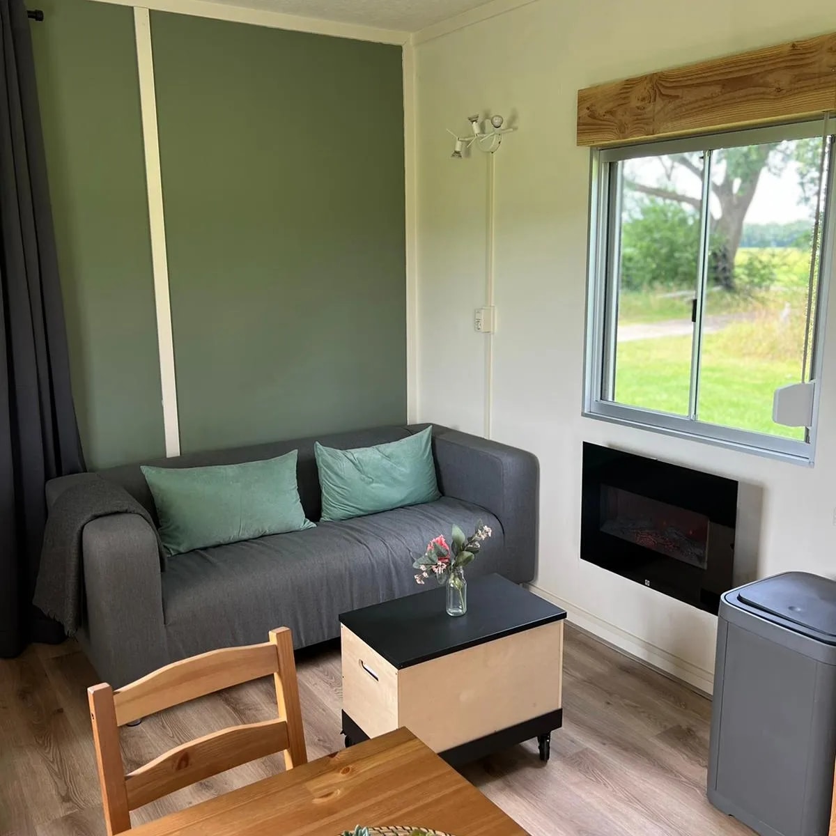 A welcoming living area features a grey sofa adorned with green cushions, positioned next to a window offering views of the surrounding fields. A small table lies in front, while a compact fireplace adds a touch of comfort. A grey cabinet is positioned nearby.