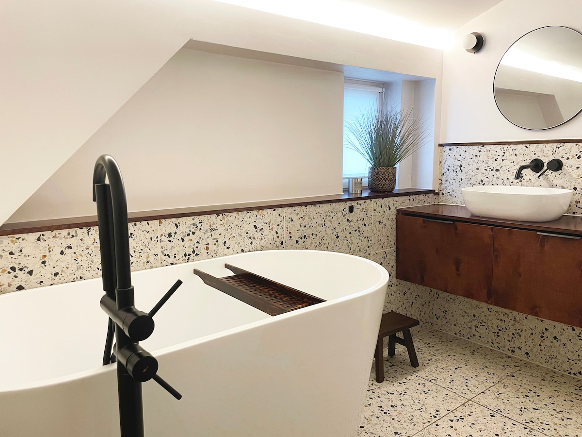 A modern bathroom features a freestanding oval tub, complemented by a round mirror above a wall-mounted sink. Textured walls with speckled materials and a wooden vanity add to the design. A small stool sits nearby, and a potted plant provides a touch of greenery.