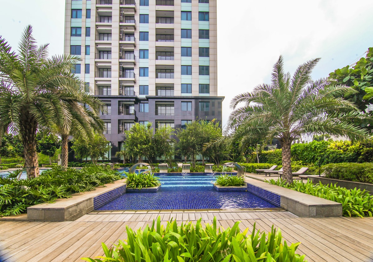 The outdoor area features a luxurious pool surrounded by lush greenery and palm trees. Modern lounge chairs are positioned by the water, and the building’s sleek architecture provides a contemporary backdrop.