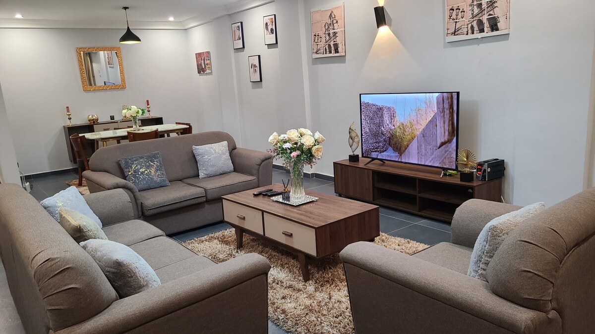 A welcoming living area features two comfortable sofas arranged around a coffee table, with decorative cushions adding softness. A television is positioned on a low cabinet opposite the seating. Fresh flowers in a vase sit on the table, and artwork adorns the walls.