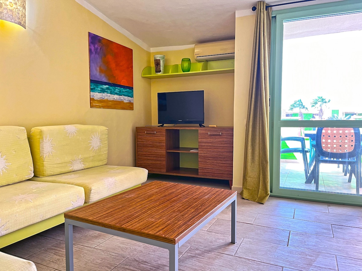 A bright living area is furnished with a light-colored sofa and a wooden coffee table. A television is mounted on a cabinet against the wall, and an abstract painting adds color to the space. Large glass doors provide access to a balcony with outdoor seating.