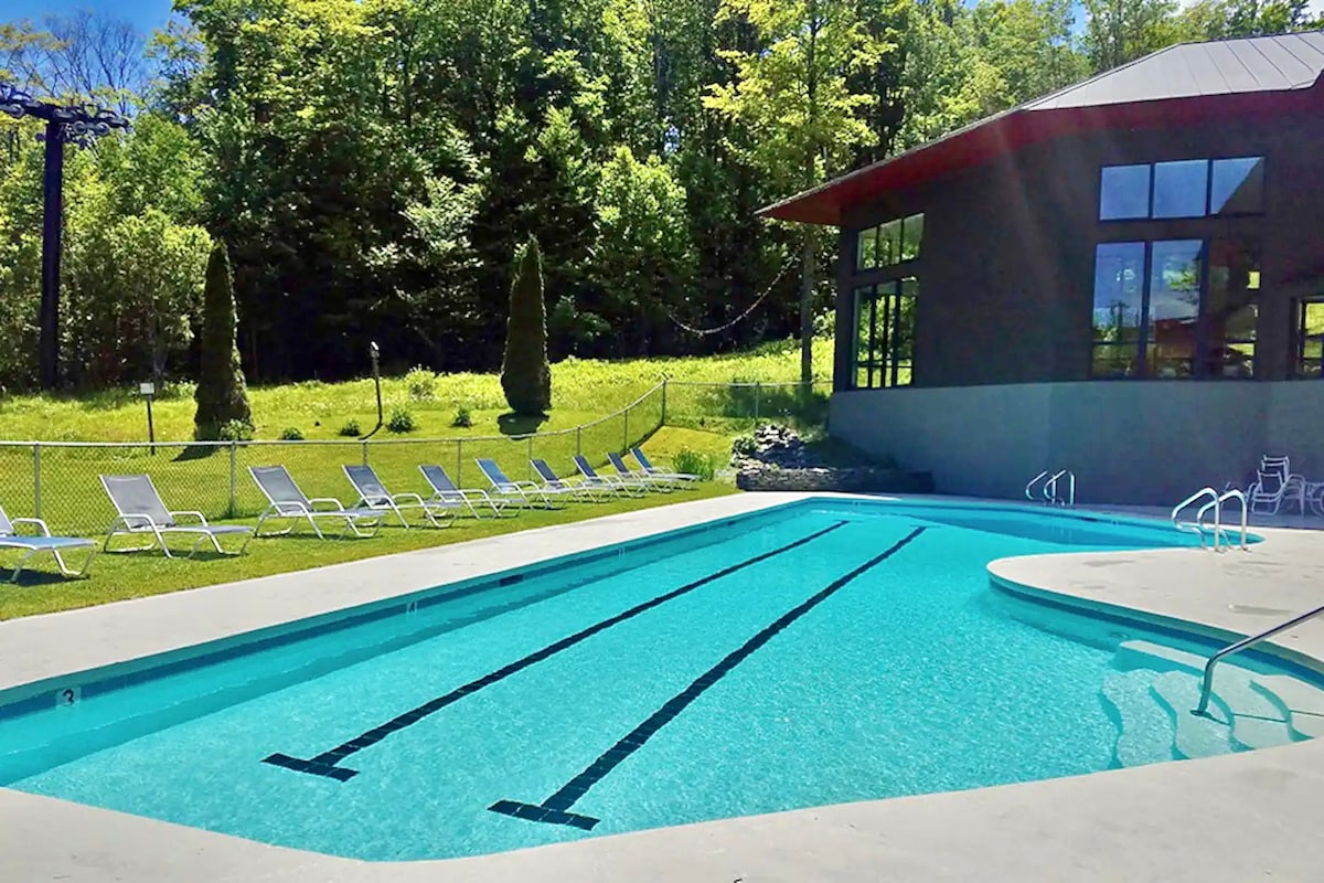 An outdoor pool is surrounded by lounge chairs, with a backdrop of lush greenery. The pool features clear, inviting water with marked lanes for swimming. A modern building with large windows is positioned nearby, complementing the tranquil outdoor environment.