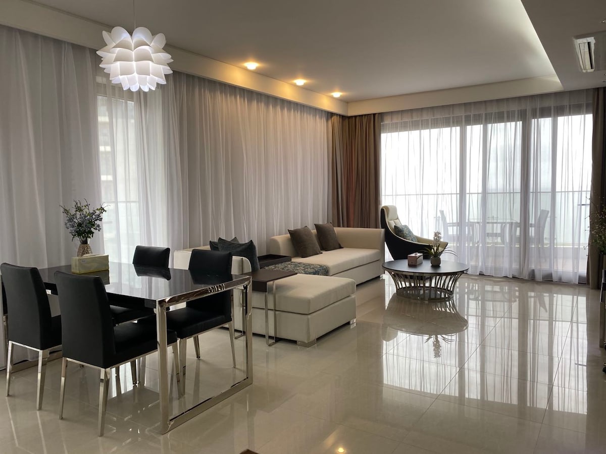An airy living space features large windows draped with sheer curtains, allowing natural light to brighten the room. A modern sofa is positioned alongside a dining table with black chairs. A tasteful light fixture hangs from the ceiling, adding a subtle touch to the room's decor.
