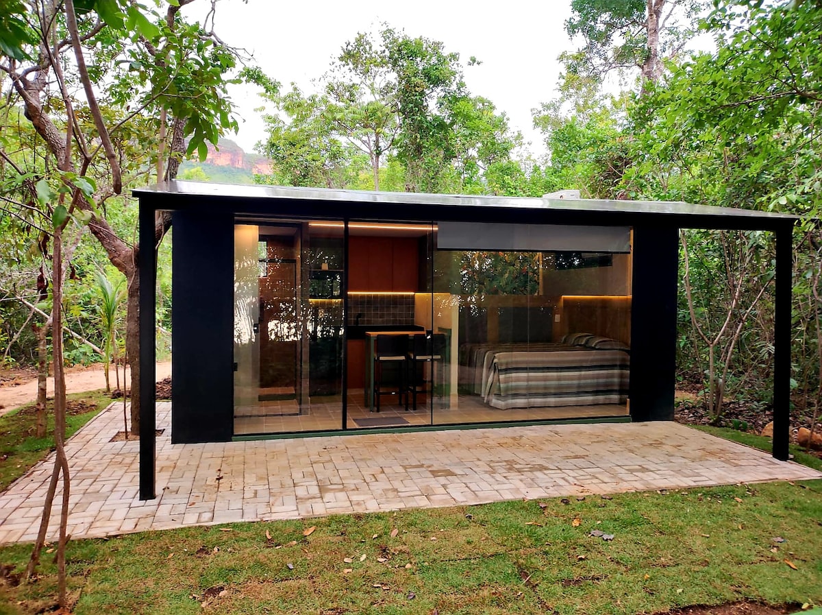 A modern container house is set in a lush natural environment. Floor-to-ceiling windows reveal an interior with a spacious bed and a dining area. The patio features a paved area surrounded by greenery, providing a peaceful connection to the outdoors.