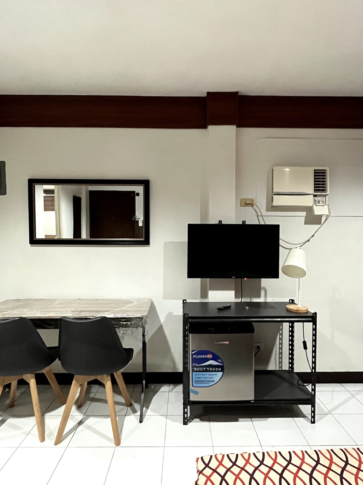 A dining area features a simple table with two chairs positioned next to a sleek TV on a rolling stand. A mirror hangs above the table, reflecting the space, while an air conditioning unit is mounted on the wall.