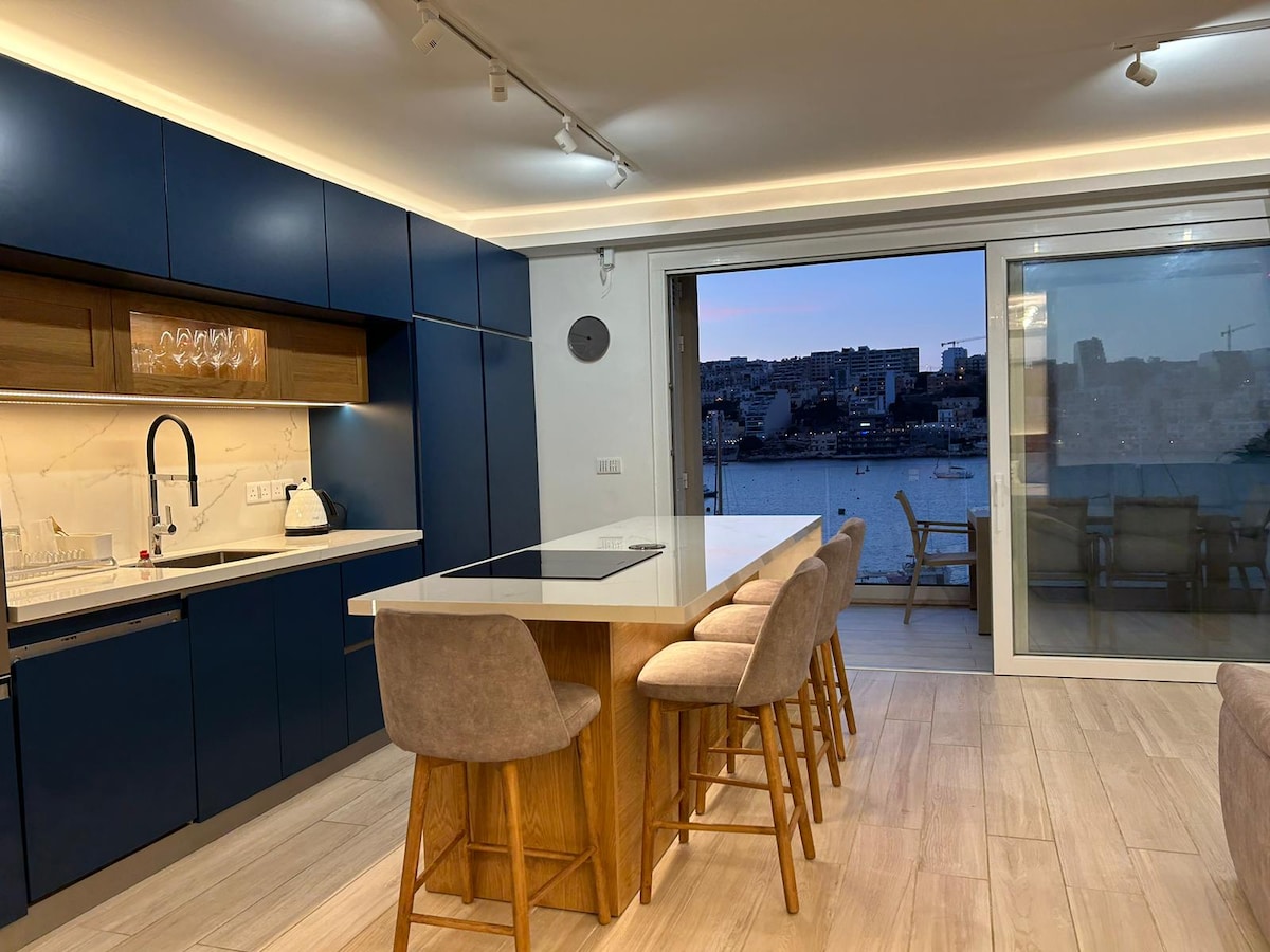 A modern kitchen features blue cabinetry complemented by wooden accents. A kitchen island with seating for four is positioned in the center. A large glass door leads to a balcony, showcasing the serene seafront view, while natural light brightens the space.