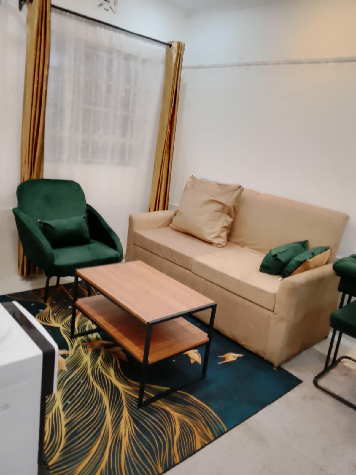 A cozy seating area features a beige sofa adorned with plush cushions. Two green chairs complement a wooden coffee table, positioned on a patterned rug. Light-colored curtains frame a window, allowing natural light to fill the space.