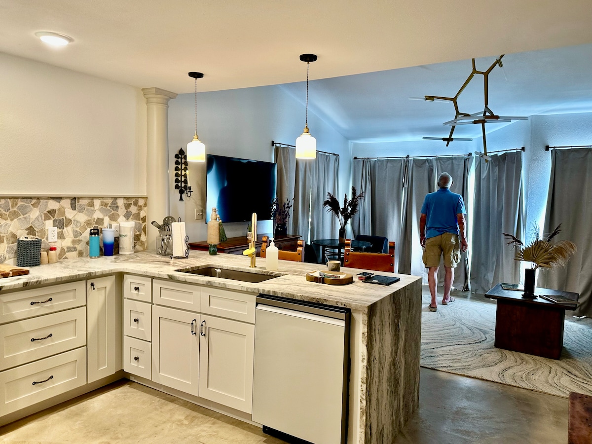 A modern kitchen with light-colored cabinetry and a marble countertop is visible. Soft lighting from pendant fixtures illuminates the space, while a dining area in the background features a large rug and draped curtains, enhancing the inviting ambiance.