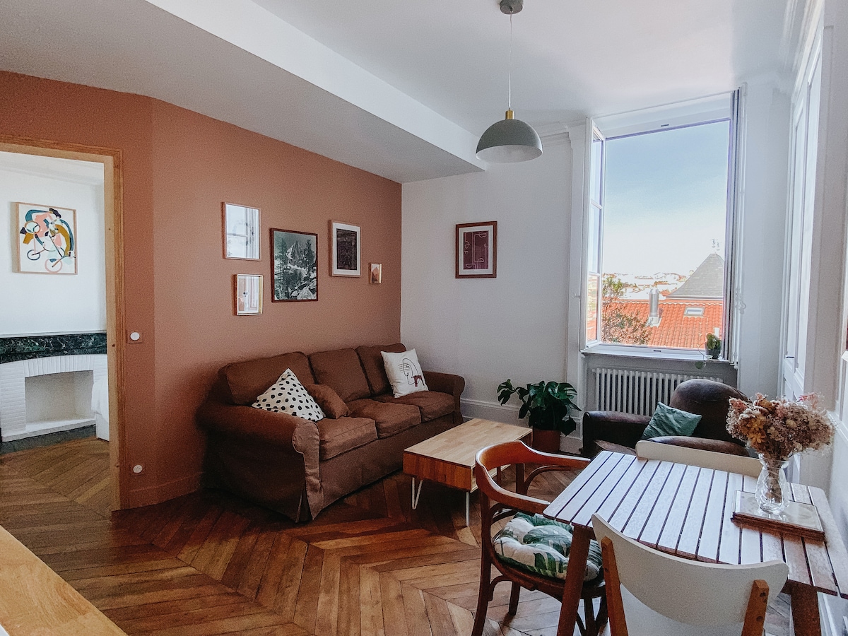 A bright living area features a brown sofa and a wooden coffee table, complemented by a small armchair. Natural light floods the space through large windows, highlighting the wooden flooring. Framed artwork adorns the walls, and a decorative vase of dried flowers adds a touch of warmth.
