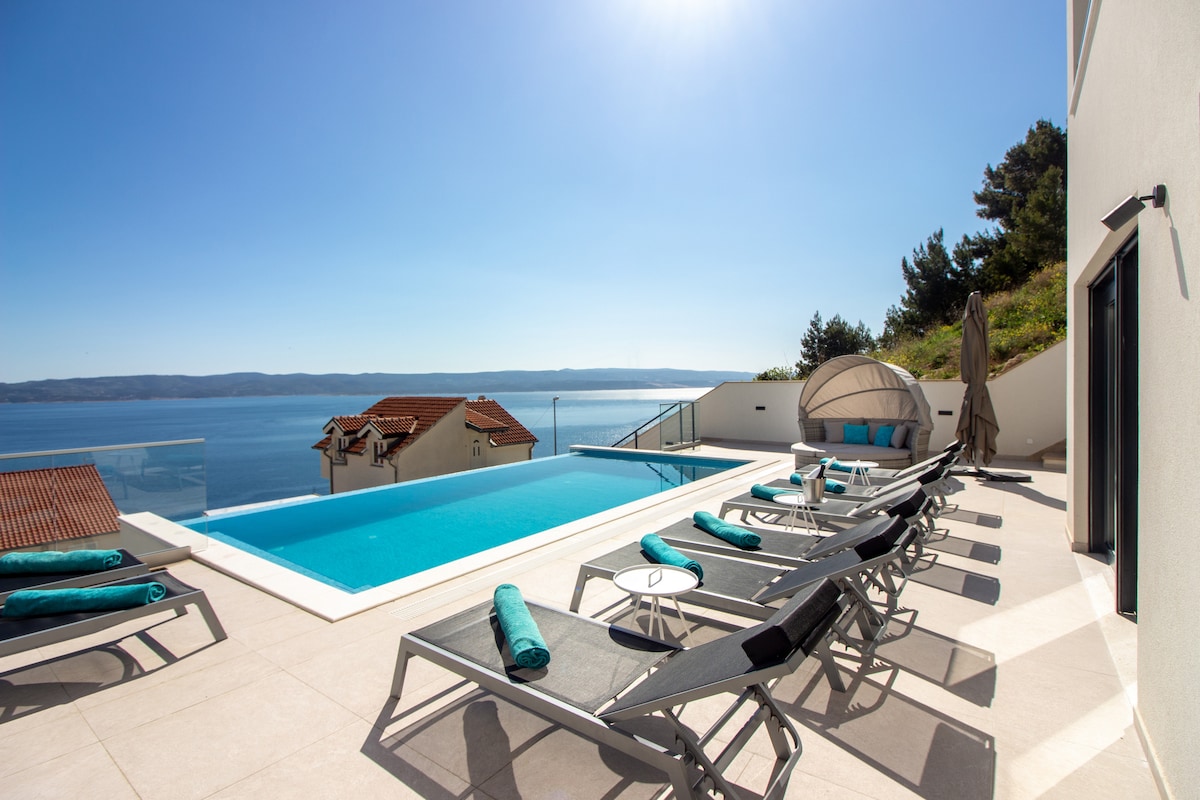 An outdoor terrace features a crystal-clear infinity swimming pool, surrounded by multiple lounge chairs with rolled towels. A shaded seating area is present, along with a minimalist table. The scenery offers expansive views of the sea and distant islands under a clear blue sky.