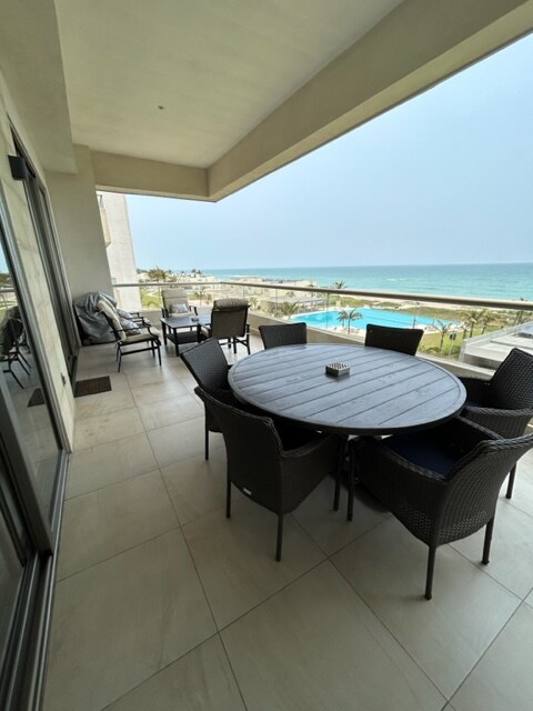 A covered outdoor terrace features a circular dining table surrounded by six wicker chairs, offering a view of the expansive ocean. The space includes a lounge area with cushioned seating, and sliding glass doors connect the terrace to the interior.