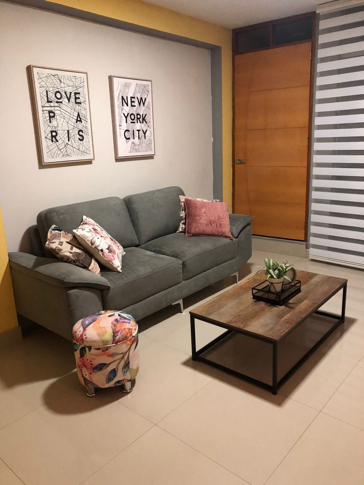 A cozy living area features a grey sofa adorned with floral-patterned cushions and a soft pink throw pillow. A wooden coffee table is positioned in front of the sofa, displaying a small plant. Two framed art pieces hang on the wall, alongside a door and light-filtering blinds.