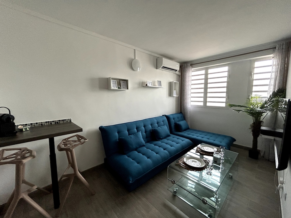 A modern living area features a plush blue sofa in a U-shape, complemented by a clear glass coffee table. Light-colored walls and large windows contribute to the bright atmosphere. A dining area with stools is positioned nearby, along with decorative shelves and a potted plant.
