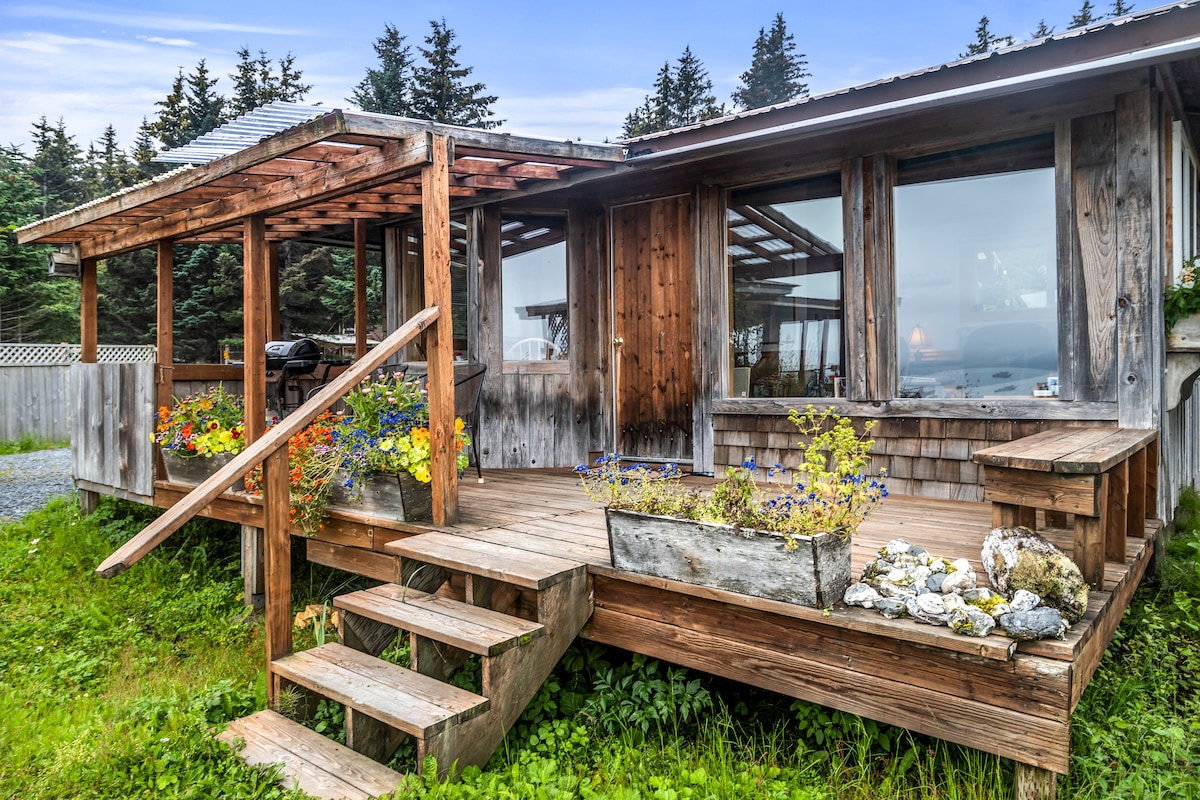 The exterior of the Seaside Cardinal cabin is presented, featuring a wooden deck with steps leading up. Colorful flower boxes are filled with blooms, adding a charming touch. Large windows allow for views of the surrounding natural landscape, while the rustic wood siding complements the tranquil setting.