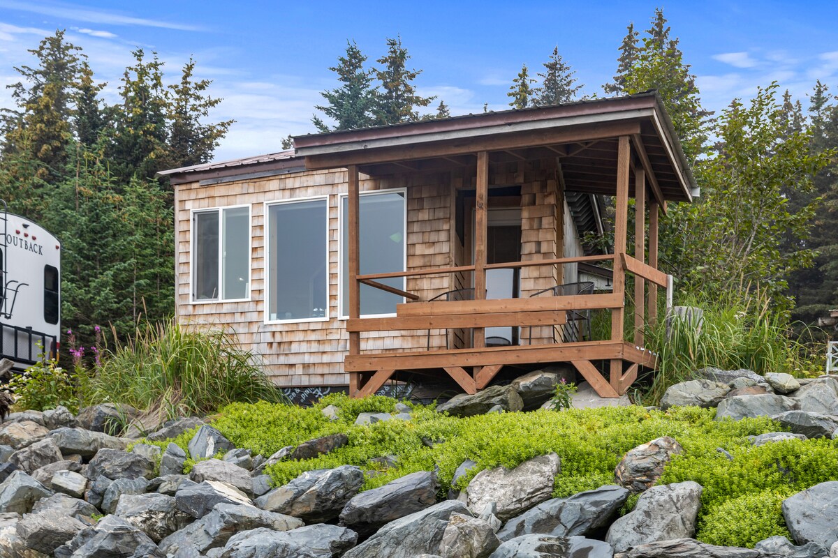 A rustic cabin is positioned on rocky terrain, surrounded by low green vegetation. Large windows provide natural light and views of the exterior, while a wooden deck extends from the front entrance, offering a welcoming space to enjoy the outdoors.