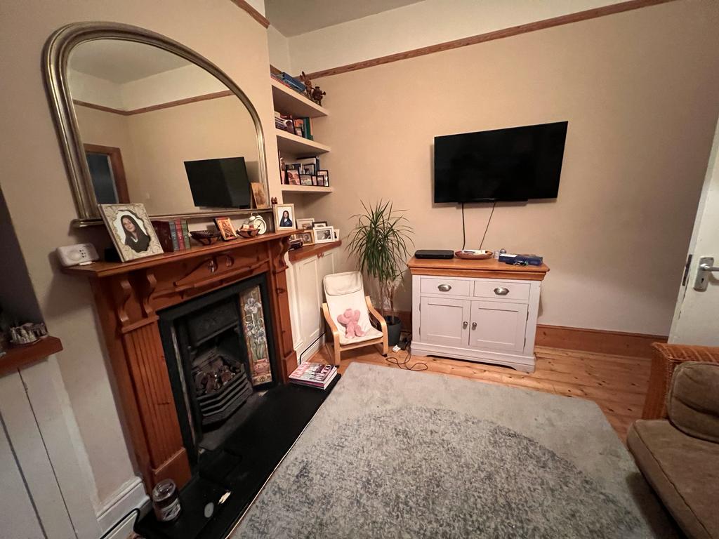A living area featuring a wooden fireplace with decorative elements and a large wall-mounted television. A light-colored rug covers the wooden floor, and a small plant adds a touch of greenery to the space. Shelving displays various photographs and decor items.