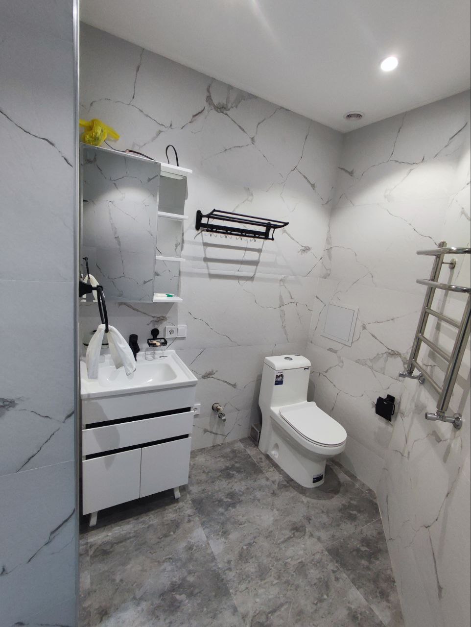 A modern bathroom is visible, featuring marble-patterned walls and grey tiled flooring. A compact sink is positioned beside a toilet, while a wall-mounted shelf and towel rack provide additional storage. Soft lighting is provided by recessed ceiling fixtures.