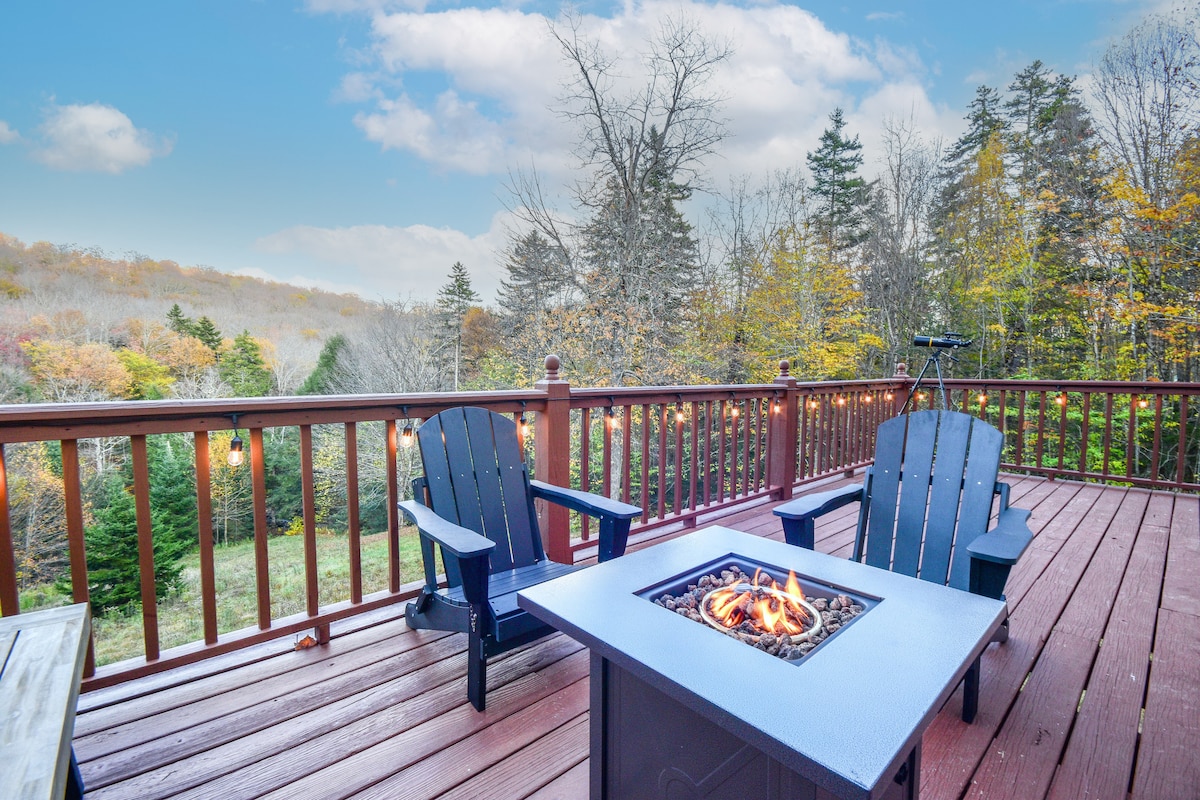 An outdoor deck is showcased with two black Adirondack chairs arranged around a square fire pit, where a warm fire burns. A telescope is positioned nearby, and string lights create a cozy ambiance, enhancing the view of the surrounding trees and autumn foliage.