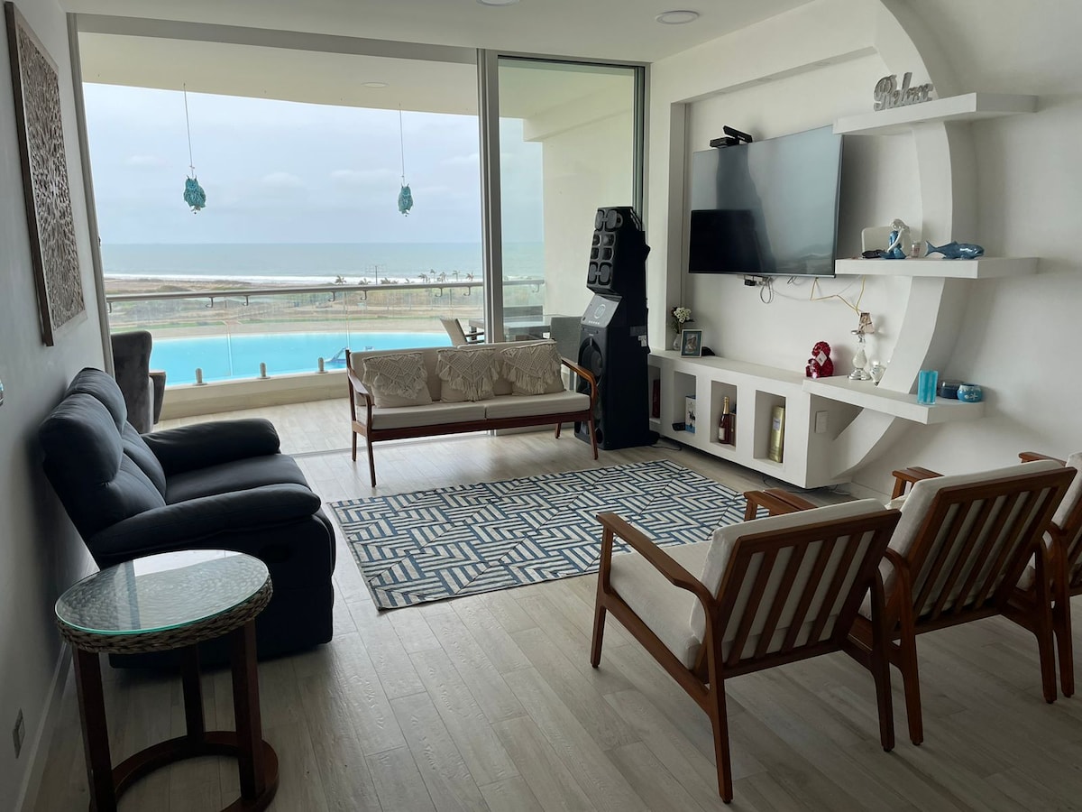 A modern living area is showcased with a comfortable dark sofa and two wooden armchairs. A patterned area rug sits beneath a circular side table, while a flat-screen TV is mounted on the wall. Large glass doors offer a view of the private beach and lagoon.