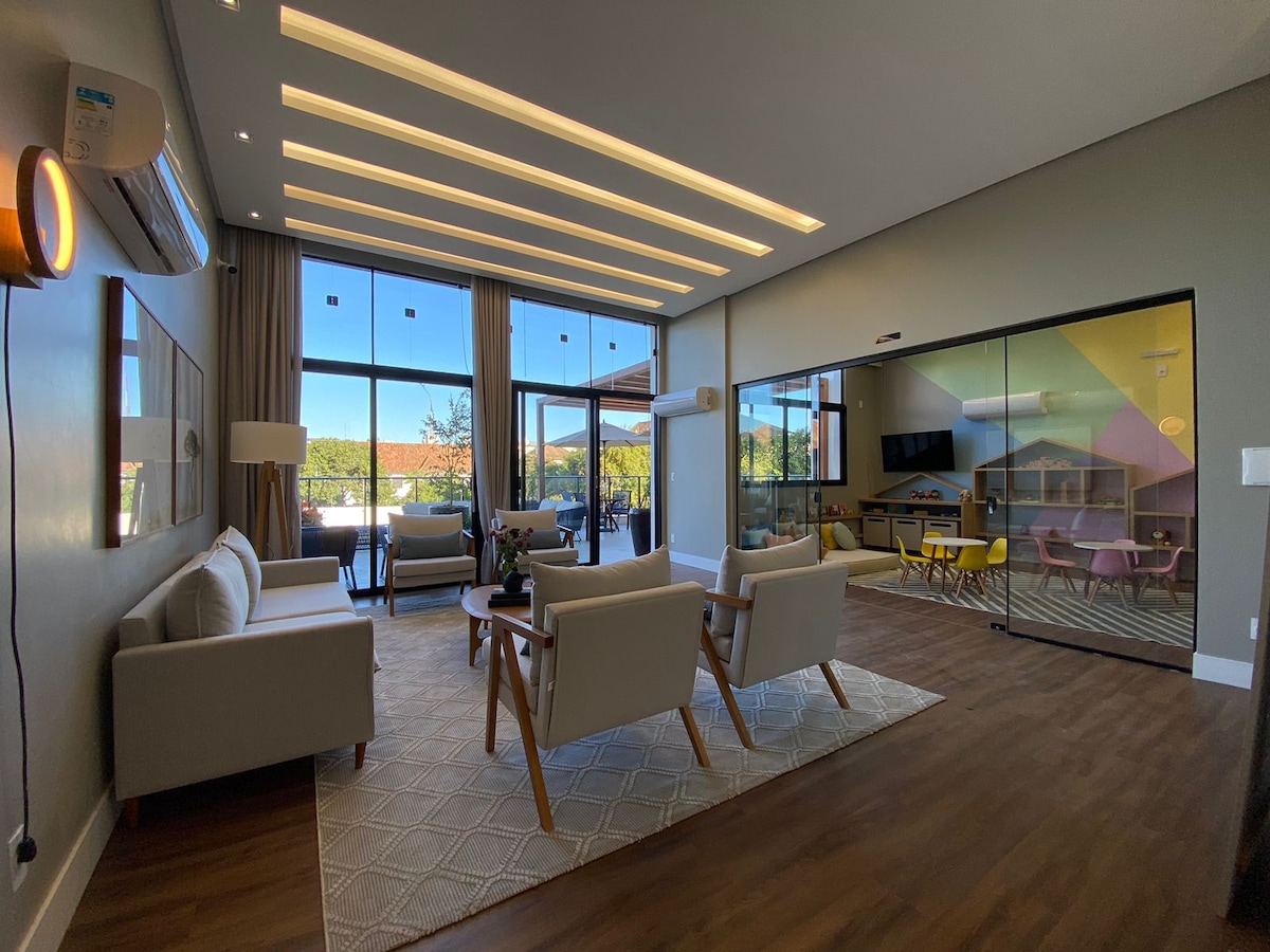 A modern living area features a light wood floor and soft-colored walls, illuminated by recessed lighting. Comfortable seating is arranged around a large area rug, with floor-to-ceiling windows providing ample natural light and a view of the outdoor space.