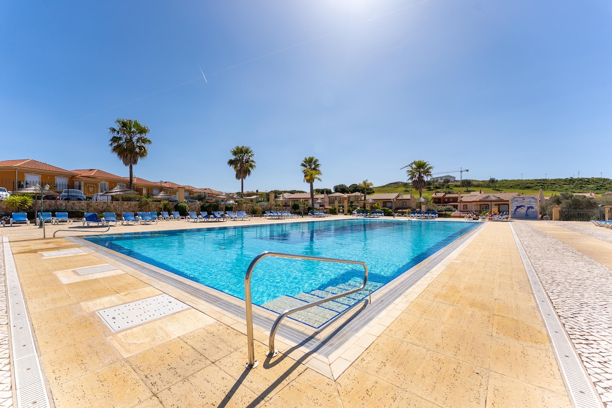 An outdoor pool area features clear blue water under a bright sky. Sun loungers lined along the pool’s edge provide ample space for relaxation. Lush palm trees surround the area, contributing to the serene atmosphere of the resort.
