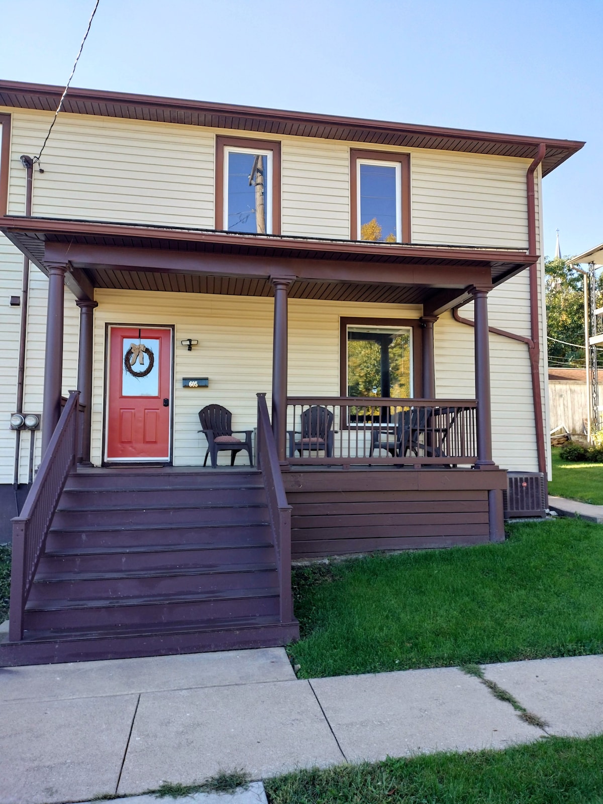 The exterior of the duplex is showcased, featuring a welcoming front porch with two chairs and a small table. The entrance boasts a vibrant red door accented by a seasonal wreath. A lawn area frames the front, with a concrete pathway leading to the steps.