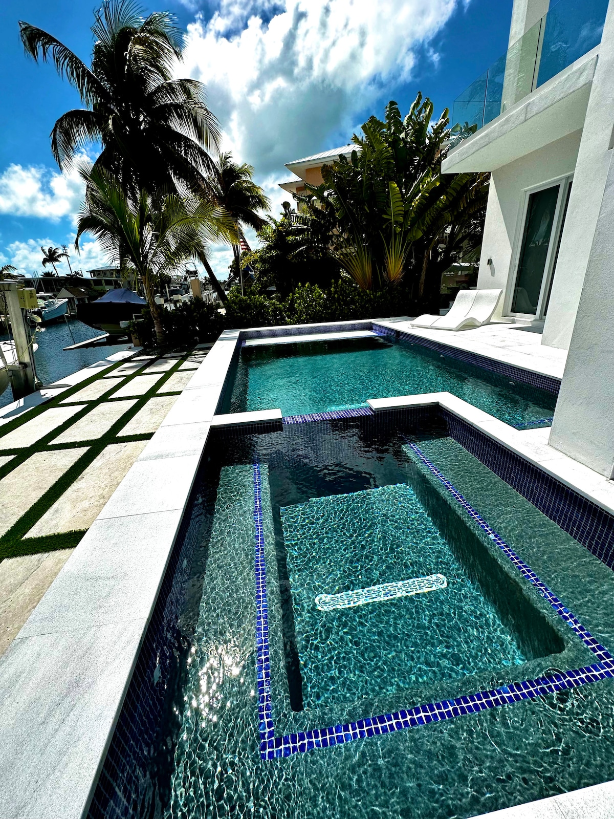 A private pool area features two connected sections, including a spacious swimming area and a jacuzzi. Surrounding palm trees and well-maintained landscaping create a serene atmosphere. Bright blue skies and fluffy clouds reflect on the water's surface, inviting relaxation.