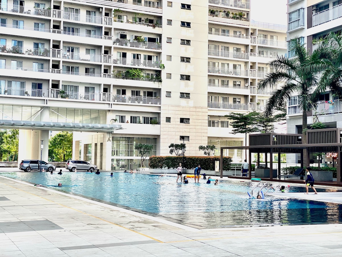 A large swimming pool is surrounded by modern buildings, providing ample space for guests to relax and socialize. Palm trees offer shading, while individuals enjoy leisure activities in the water. Lounge areas are visible along the poolside, enhancing the outdoor experience.
