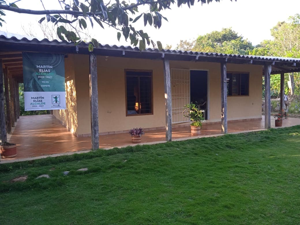 A single-story building features a covered porch with wooden posts, bordered by lush green grass. Large windows provide views of the surrounding nature, and potted plants add a touch of greenery to the entrance.