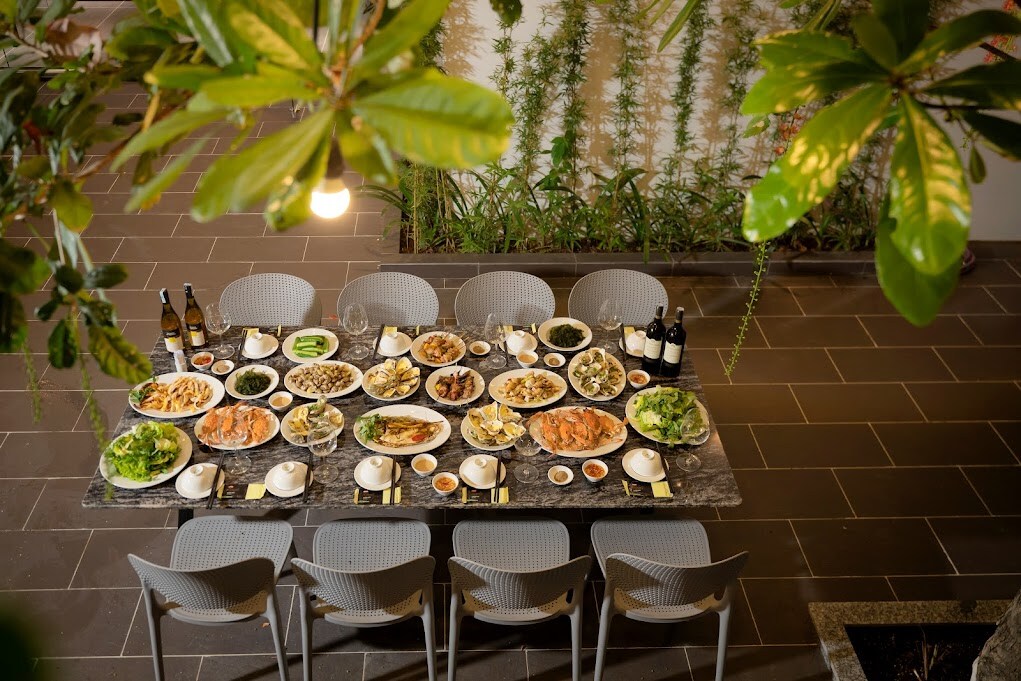 A spacious outdoor dining area features a long table set for a meal. Various dishes are arranged along the table, accompanied by glasses and bottles of wine. Green foliage surrounds the space, providing a natural backdrop.
