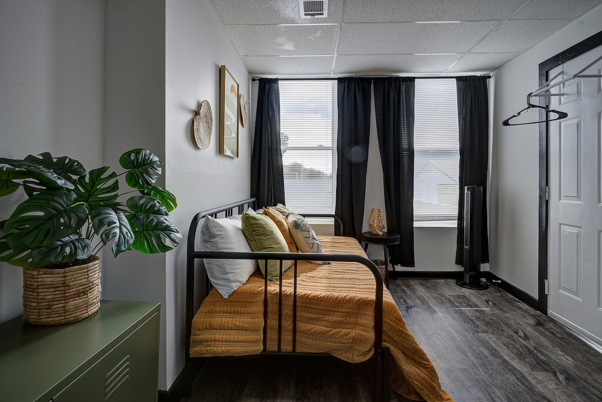The room features a trundle bed adorned with beige and green bedding. Natural light fills the space through two large windows with black curtains, while a plant and decorative wall art add a touch of warmth. A closet and a fan are positioned near the door.