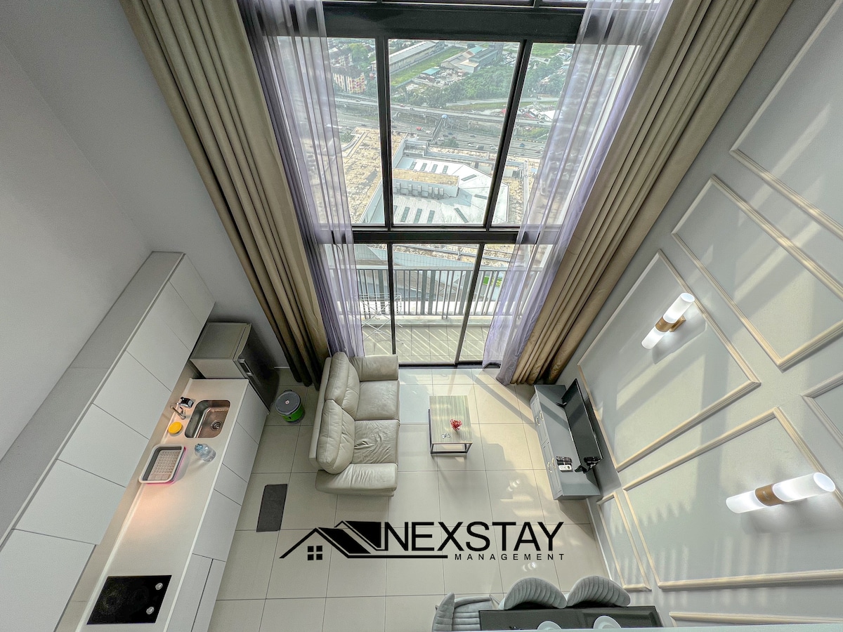 An aerial view captures the spacious living area with a light-colored sofa and a small coffee table, framed by large floor-to-ceiling windows that provide natural light. The kitchen area showcases modern cabinetry and appliances, accentuated by neutral tones and elegant decor.