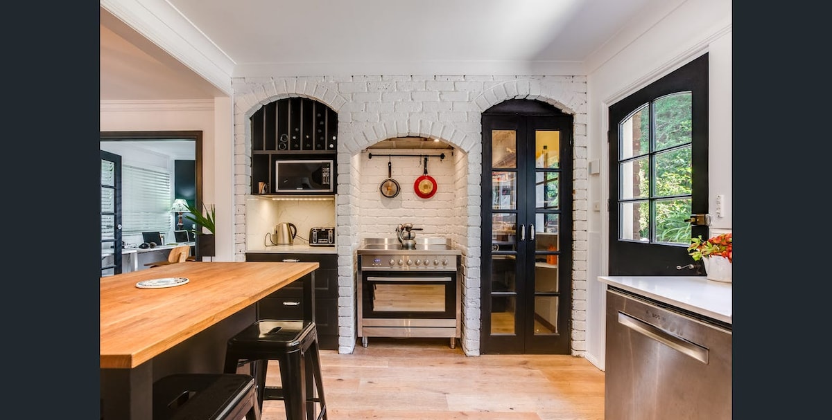 The kitchen features white brick walls with a wooden countertop and black stools. Stainless steel appliances, including a range and microwave, are complemented by dark cabinetry. An elegant glass door leads to an adjoining space, while decorative hanging cookware adds a functional touch.