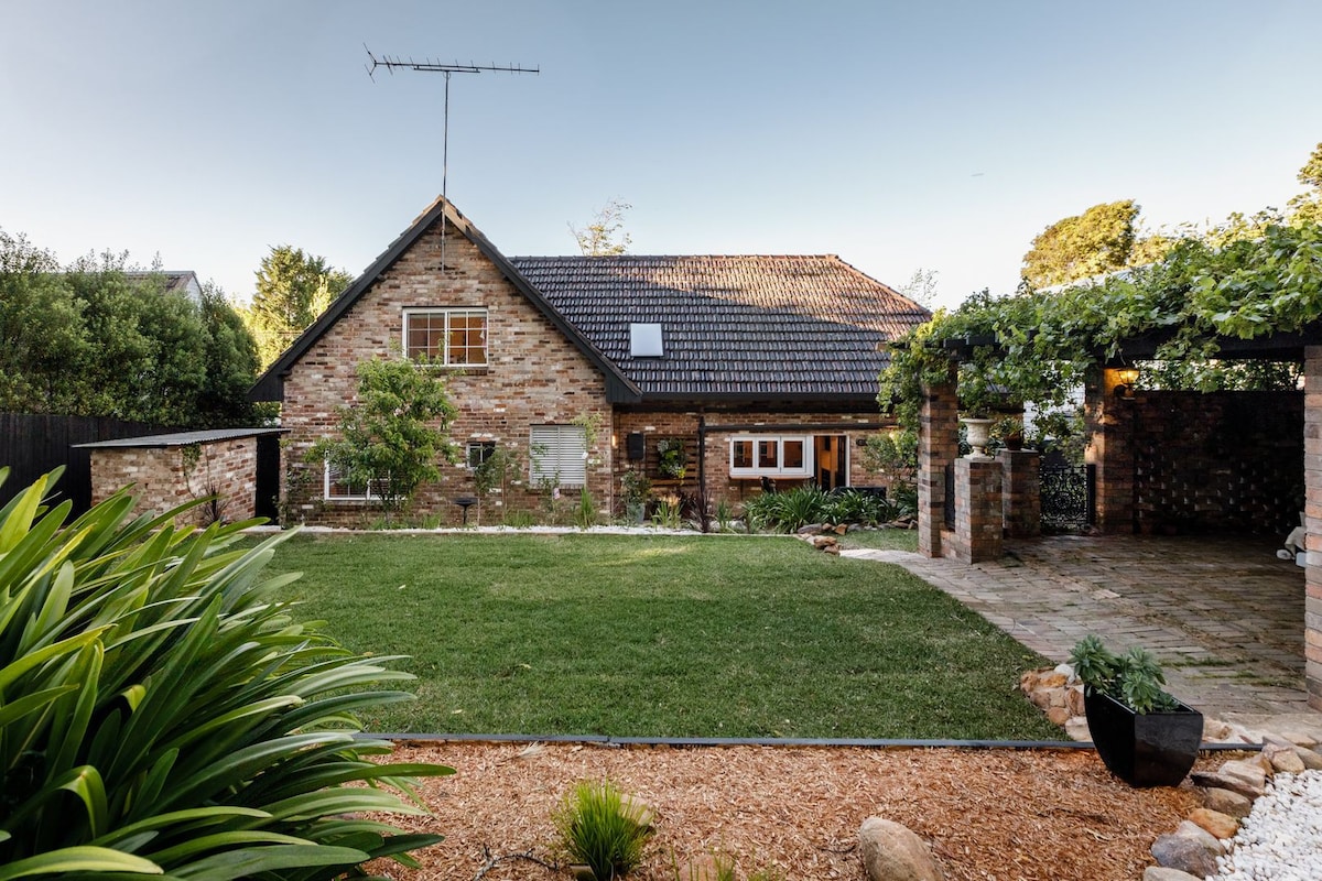 A charming brick cottage is nestled within a well-manicured lawn, framed by lush greenery. The building features a steeply pitched roof and multiple windows, allowing natural light to fill the interior. A stone-paved outdoor area and trellised vines add to the inviting ambiance.