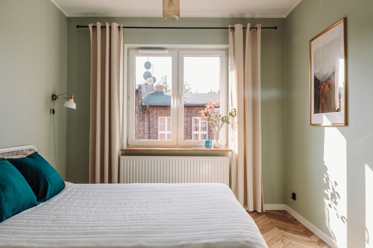 A well-lit bedroom is furnished with a spacious bed with teal accent pillows. Large windows are framed with light curtains, allowing natural light to illuminate the room. A decorative photo and a small flower arrangement add a touch of detail to the neutral-colored walls.