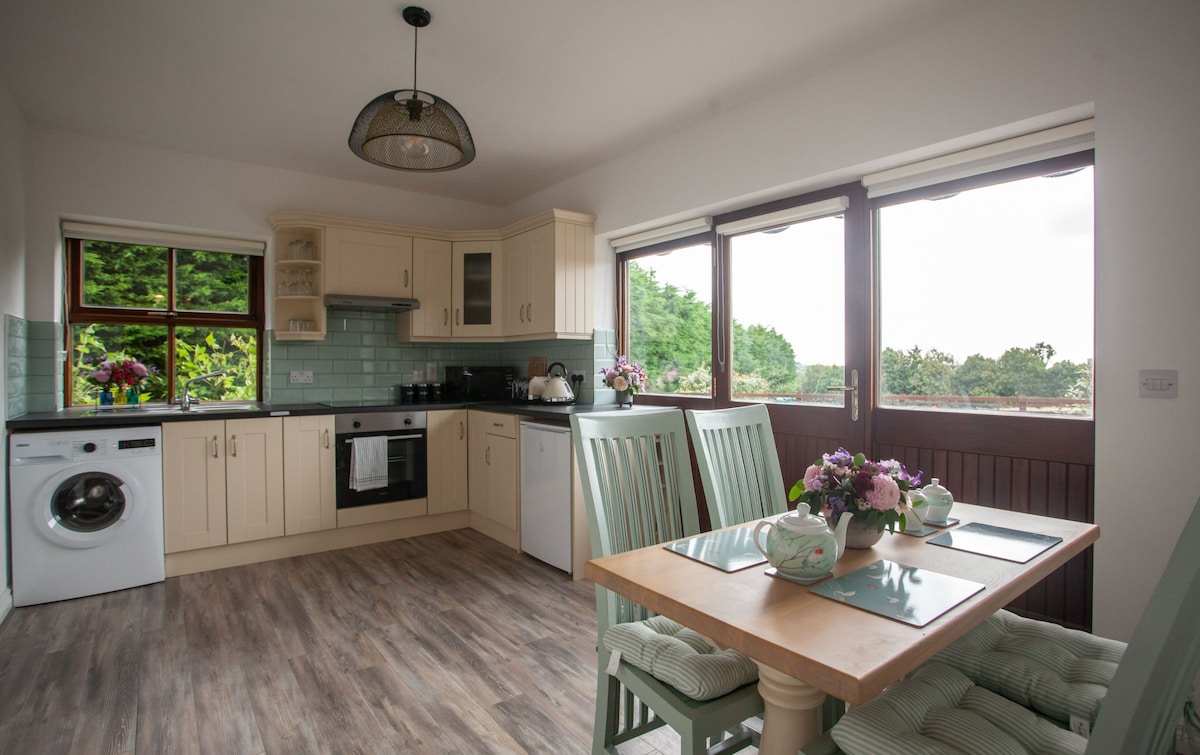 A bright kitchen area is filled with natural light from multiple windows, showcasing a dining table set for two. Subtle mint-colored accents are present in the seating and cabinetry. A washing machine and modern appliances are visible, enhancing functionality.