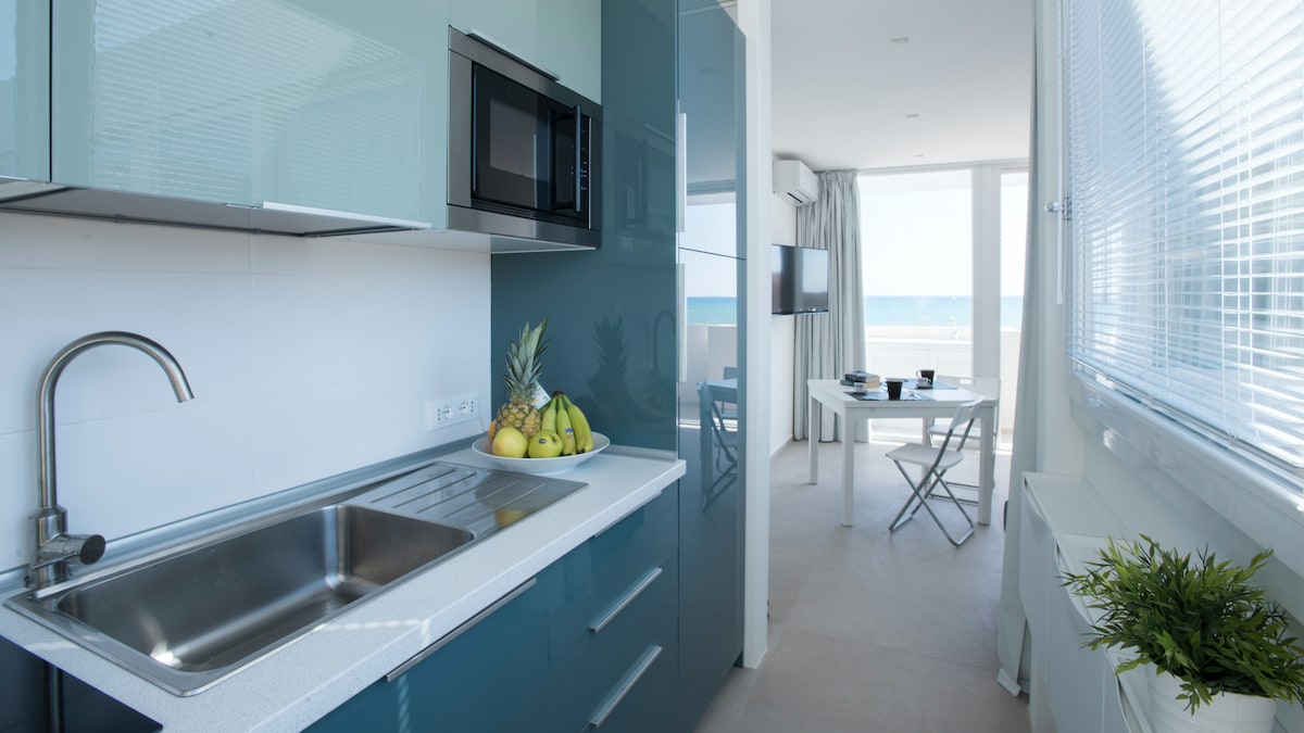 A modern kitchen area features sleek blue cabinetry and a stainless steel sink. Natural light floods the space through large windows, with a view of the sea visible beyond. A small dining table is present, accompanied by chairs, and fresh fruit is displayed on the counter.