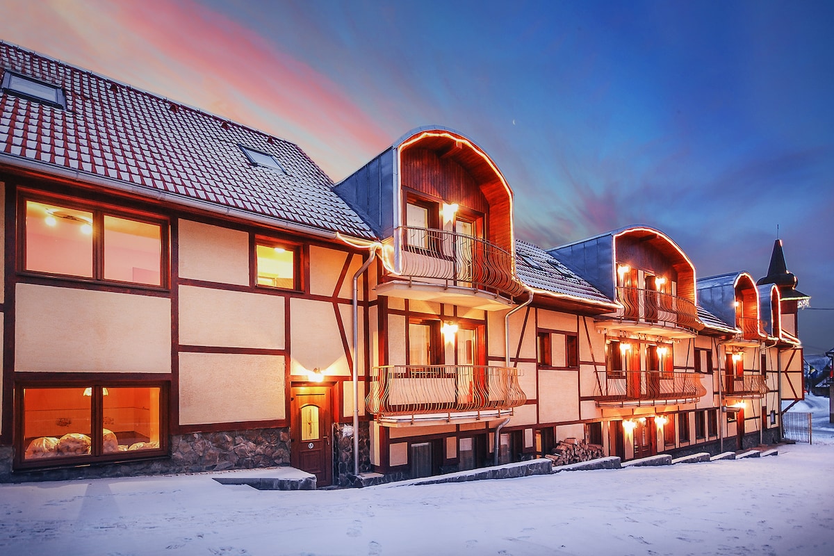 A modern apartment building is depicted, showcasing multiple levels with curved balconies. Soft evening light highlights the architectural details and the snow-covered ground. Warm illumination from windows suggests a cozy interior, inviting guests to explore the space.