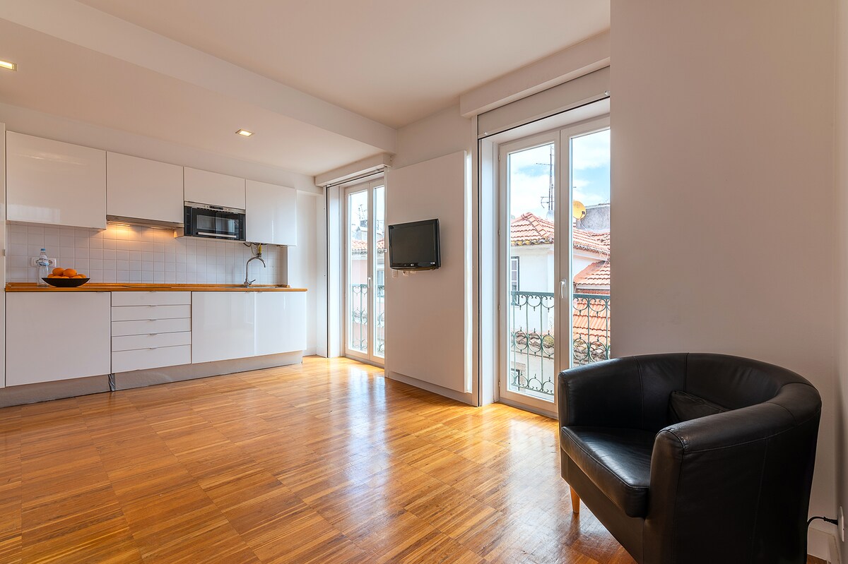 An open living space is showcased, featuring large windows allowing natural light to fill the area. A modern kitchen with sleek cabinets and a microwave is adjacent to a dining space. A comfortable armchair is positioned close to the windows, enhancing the ambiance.