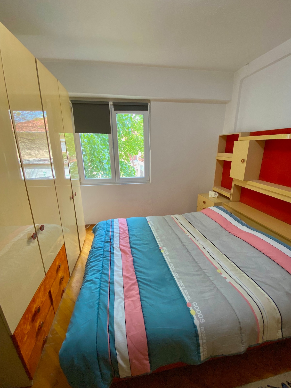 A cozy bedroom is presented with a striped bedspread covering a spacious bed. A light-colored wardrobe and shelving unit are visible, complemented by a red accent wall. Natural light enters through a window, contributing to the room's ambiance.