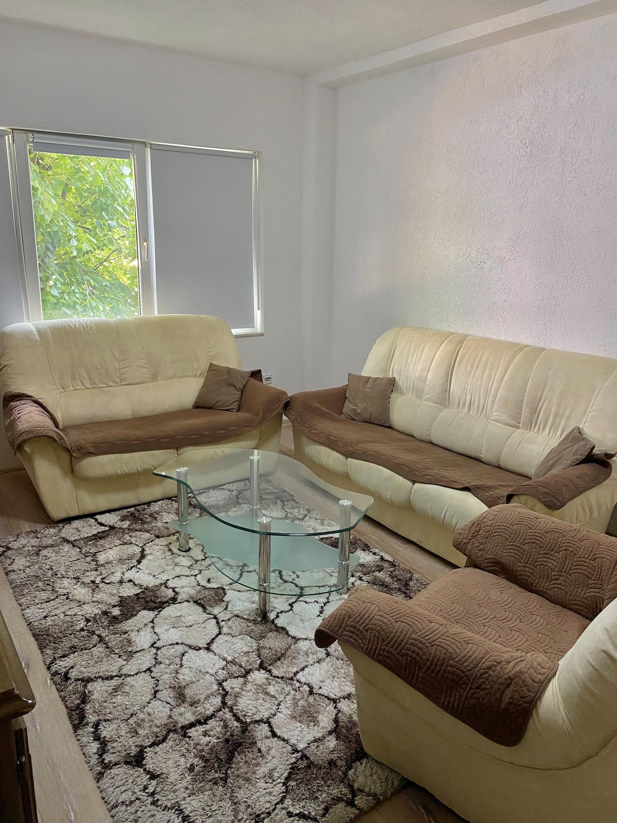 A cozy seating area is presented with two cream-colored sofas, complemented by decorative brown throws. A glass coffee table sits centrally on a textured area rug, with natural light filtering through the nearby window, enhancing the inviting ambiance.